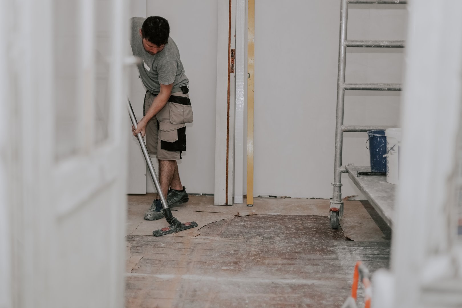 Young caucasian brunette man in work clothes on the left is vacuuming the floor with a construction vacuum cleaner in a room in an old house being renovated, side view close-up with selective focus.Construction work concept.
