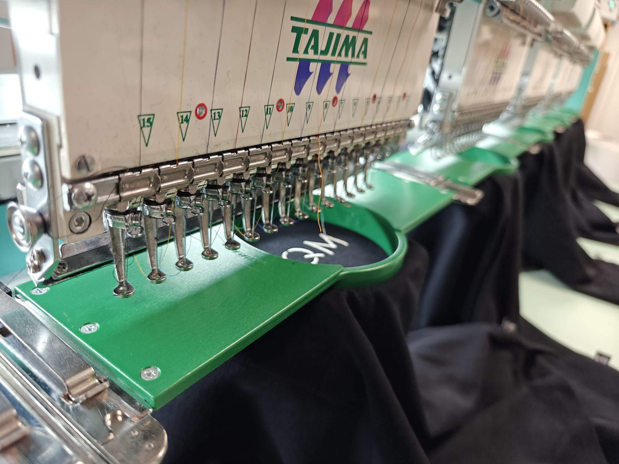 Close-up of a Tajima embroidery machine stitching a design on black fabric, showing multiple needles and threads.