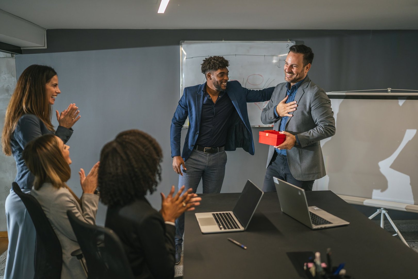 Diverse business team celebrating professional success and appreciating an employee receiving a gift box during a corporate meeting in a modern office environment