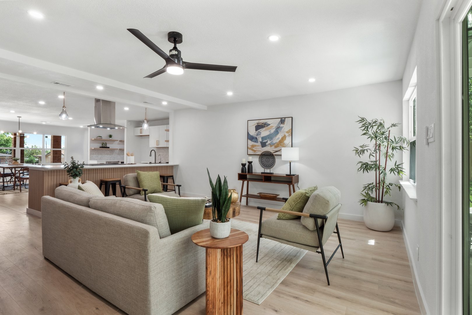 Modern living room with neutral decor, green accents, and open kitchen. Features ceiling fan, plants, and artwork.