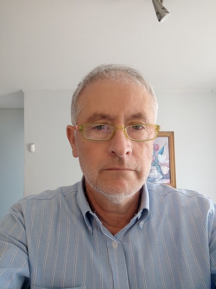 Older man with glasses and a beard, wearing a striped shirt, standing in a room with a framed picture on the wall.