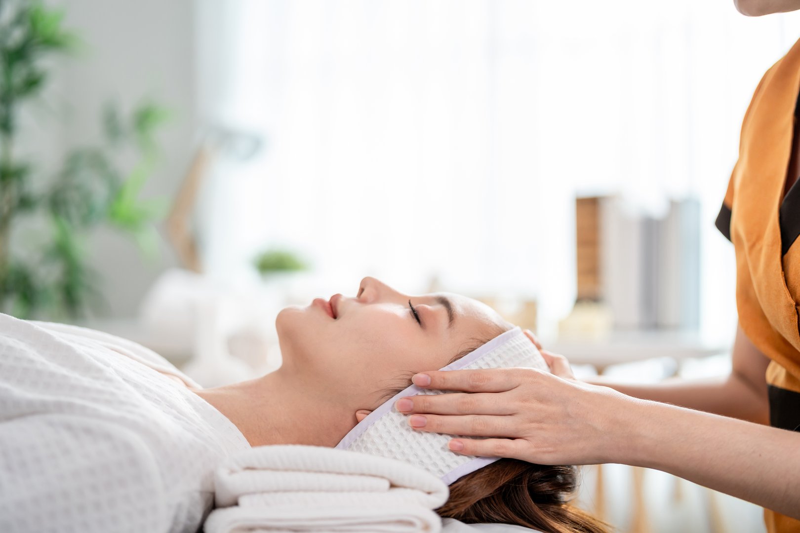 Asian young woman getting head and facial massage treatment at beauty spa. Attractive girl lying down on massage bed, getting relaxing physiotherapy service from masseuse for body care in spa clinic.