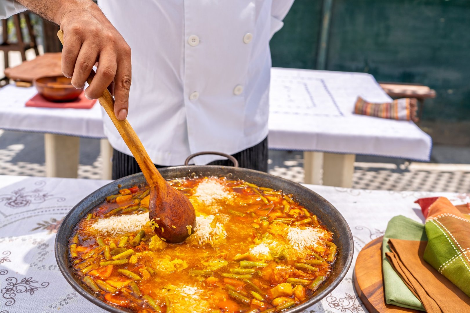 Chef preparing a delicious paella outdoors, adding rice to a large pan filled with vegetables and seafood