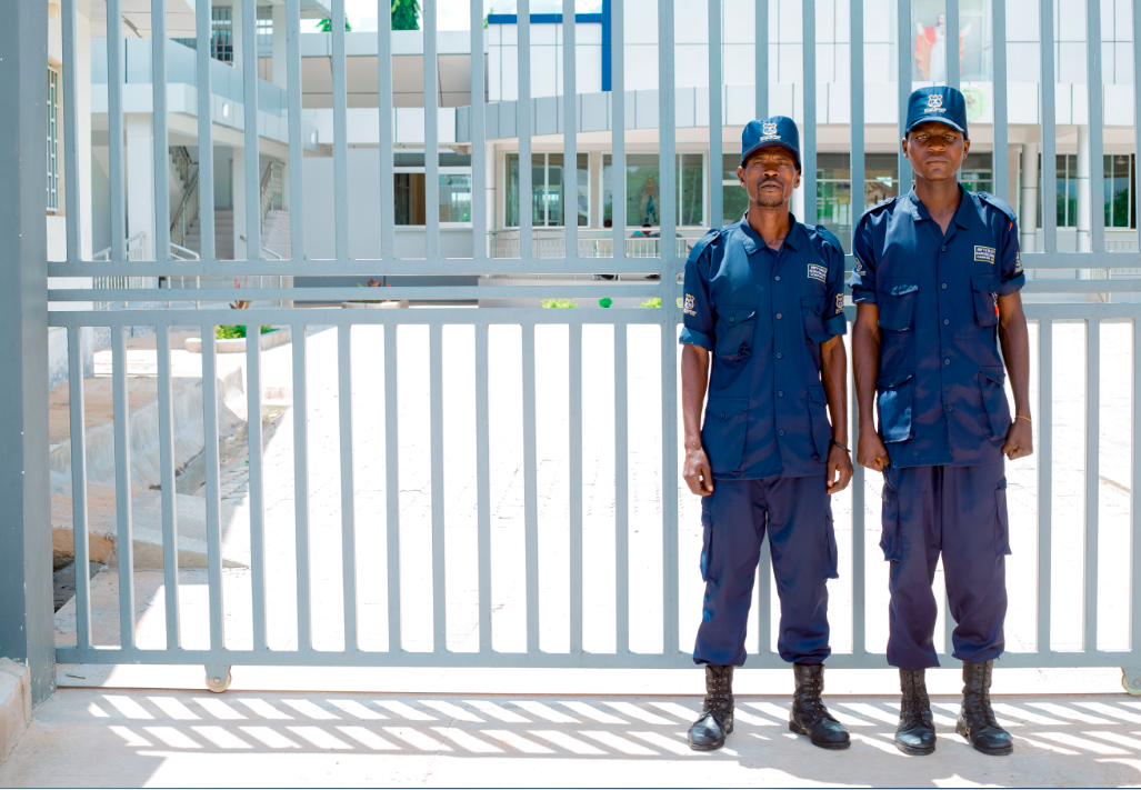 Two uniformed security guards standing in front of a gated entrance on a sunny day.