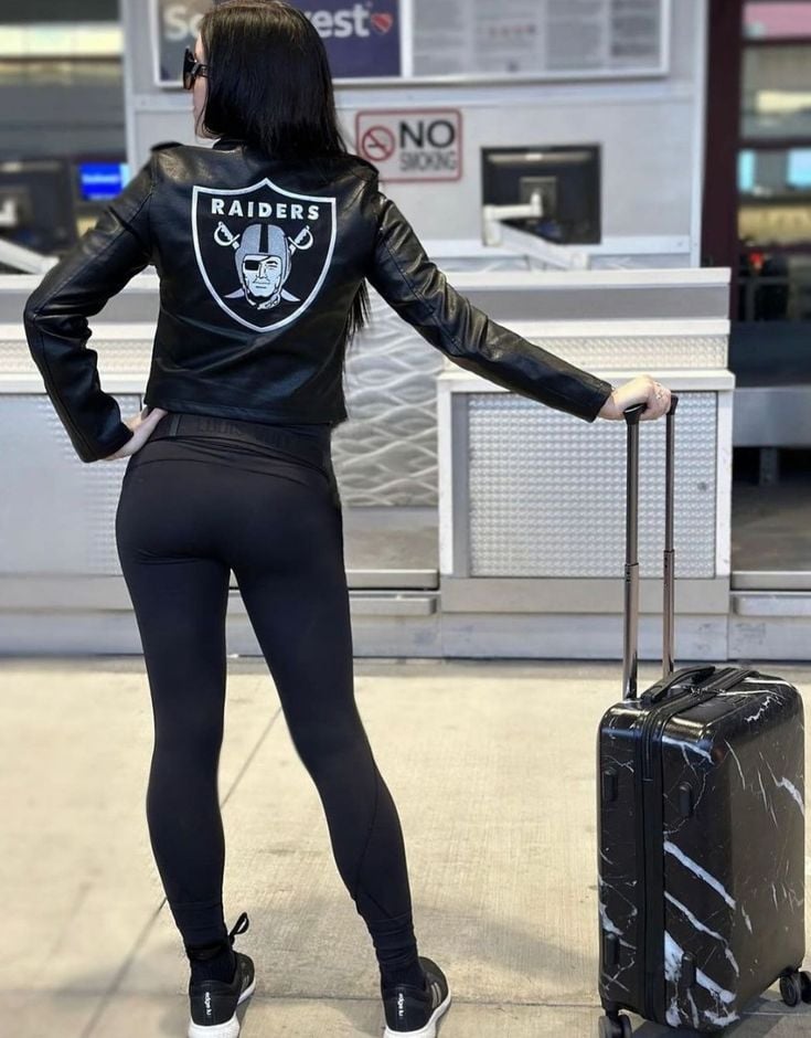 Person in a Raiders leather jacket stands with a black suitcase outside, near a no smoking sign.