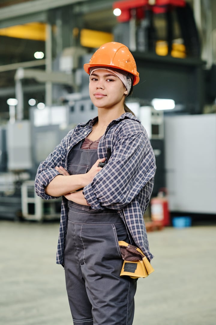 Portrait of female engineer standing with crossed arms in industrial factory environment Wearing hard hat and plaid shirt, embodying confidence and professionalism
