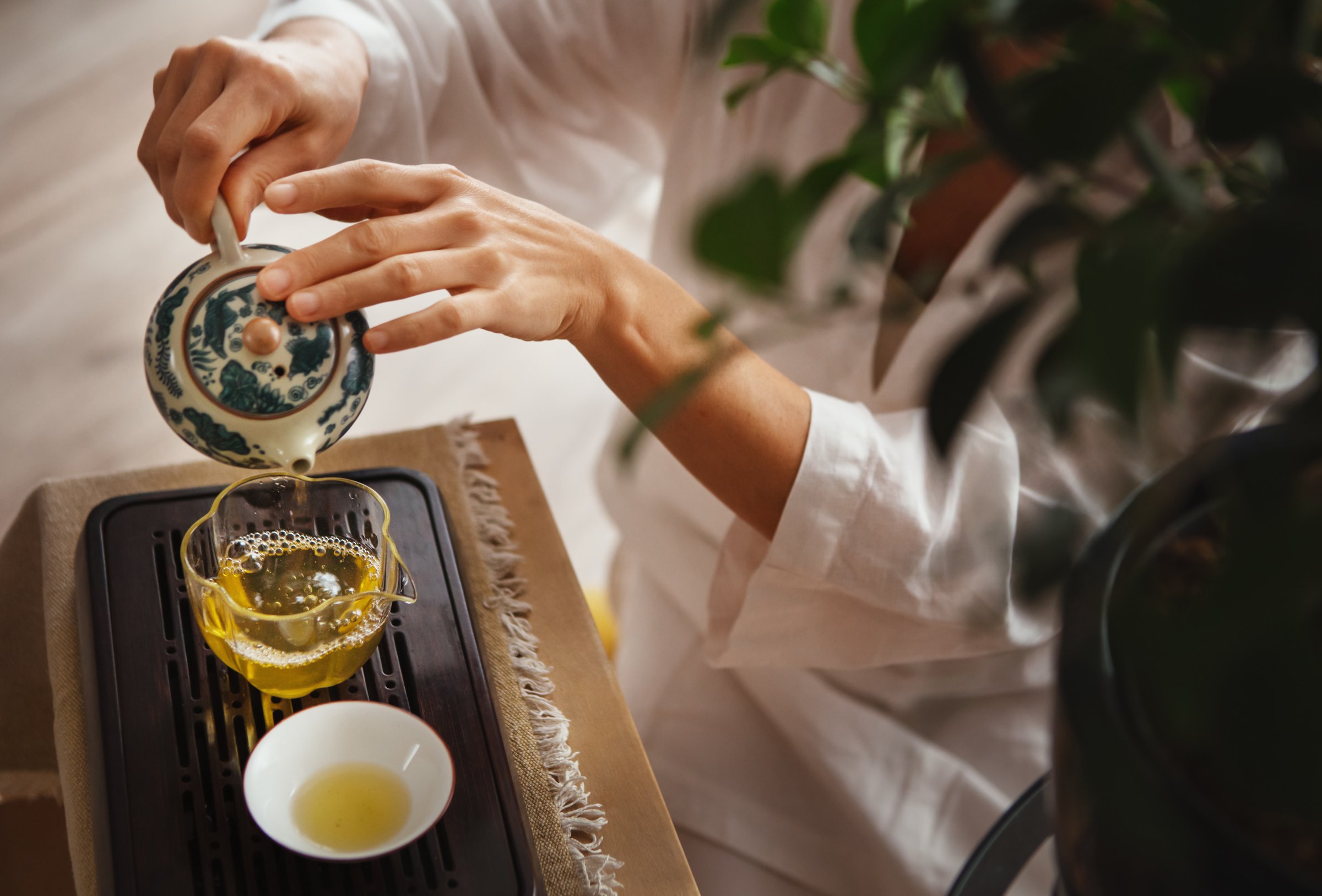 Female hands pouring tea from patterned clay teapot into clear glass pitcher, creating serene tea ritual. Soft light, wooden tea tray and greenery evoke calming mindful chenese ceremony atmosphere.