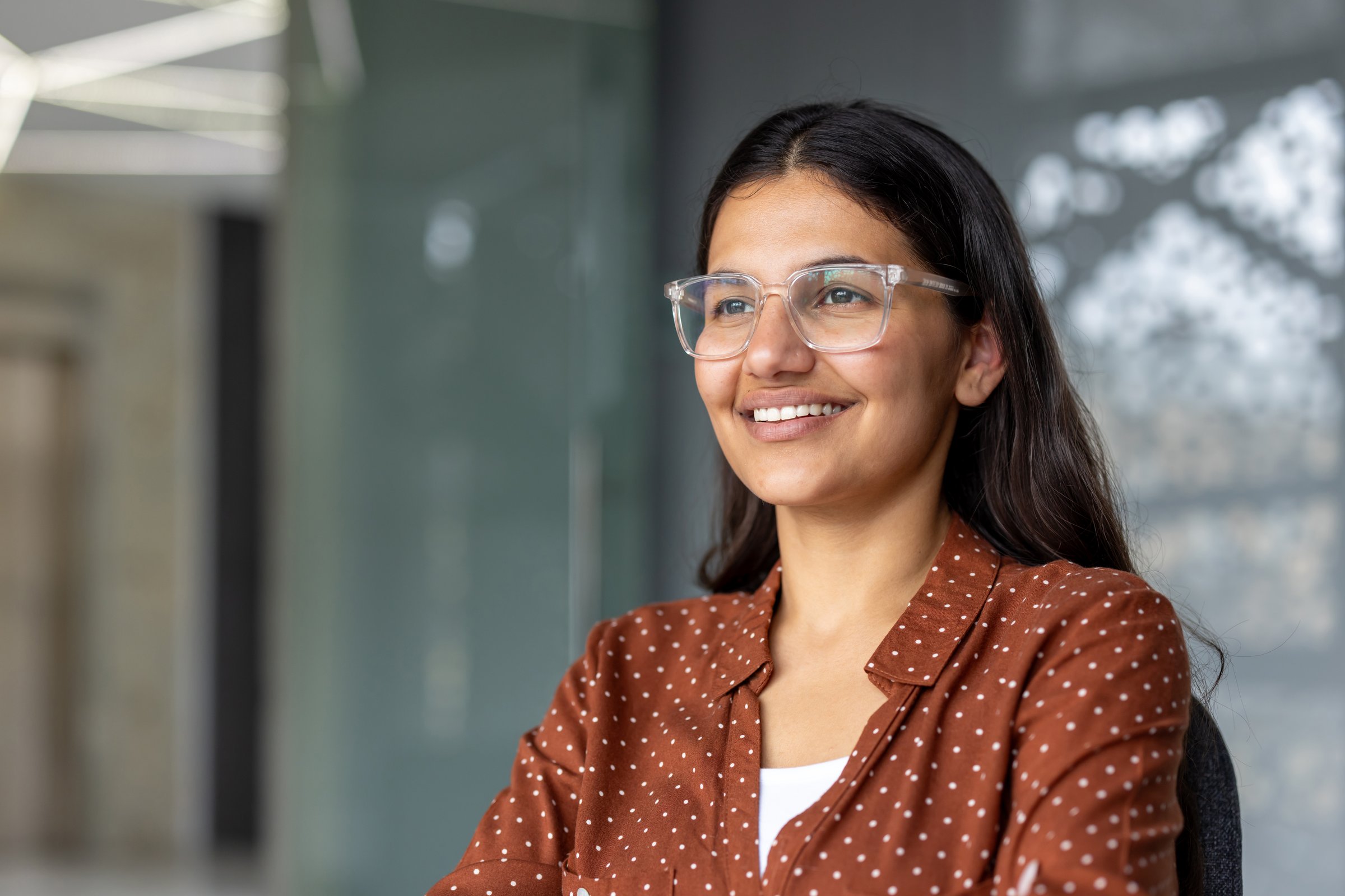 Young indian woman wearing clear glasses and a patterned shirt, smiling confidently while looking away in a modern office environment, symbolizing success, inspiration, and positive business outlook