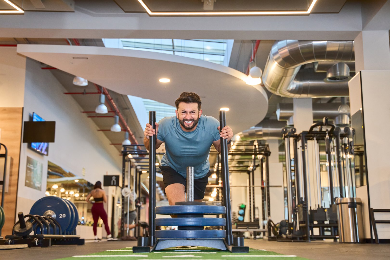 Energetic man pushing a weighted sled on turf in a contemporary gym. Fitness equipment and industrial design elements in the background