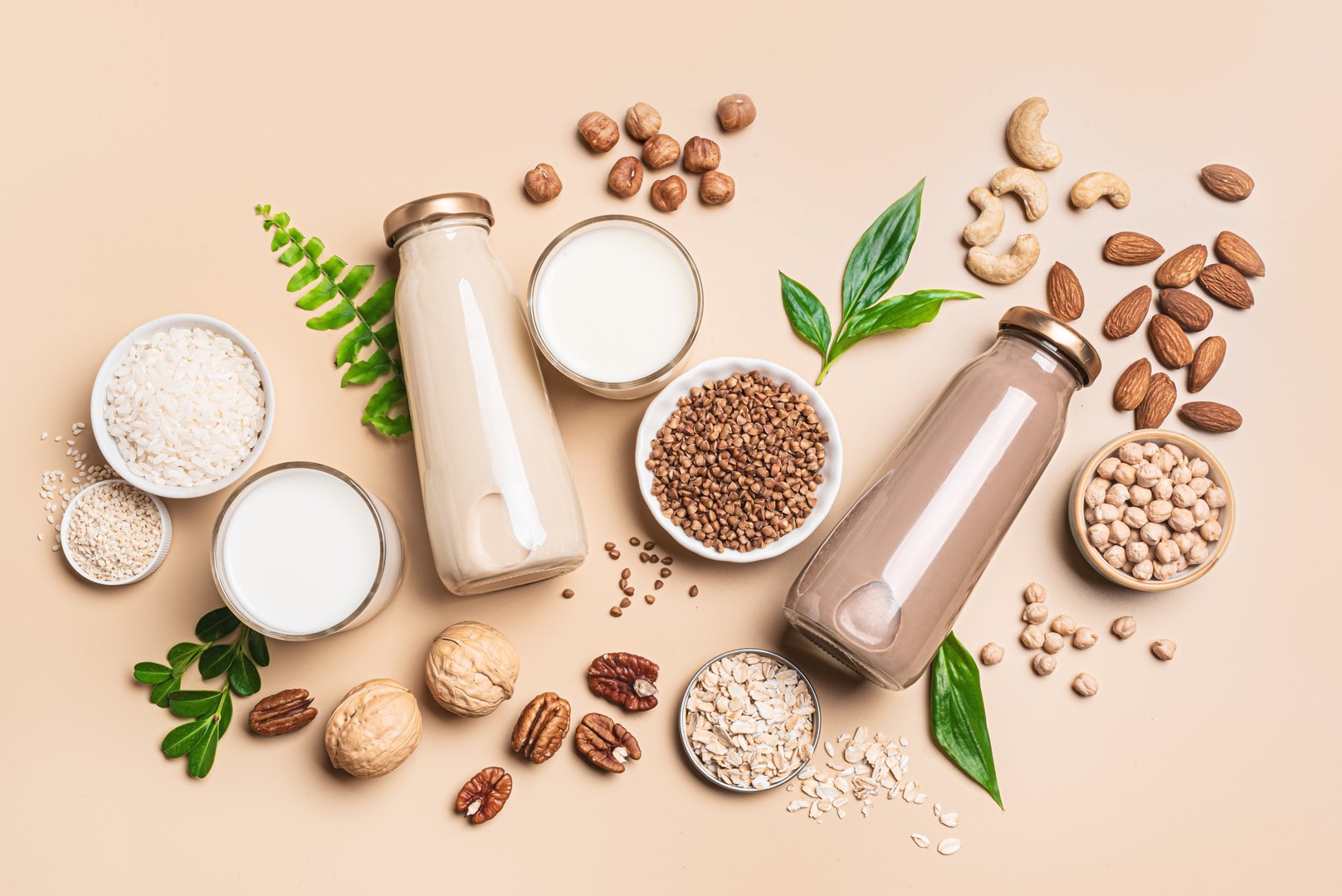 Vegan plant based milk bottles, ingredients and green leaves. Non dairy, lactose free milk with nuts and grains on neutral beige background, flat lay.  Substitute drink, healthy eating.