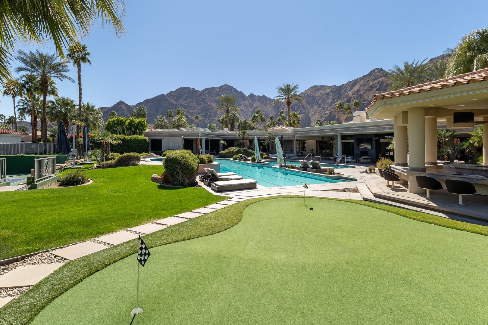 Putting green and swimming pool with outdoor bar and a mountain backdrop with palm trees at a upscale, luxury estate in Indian Wells, California.