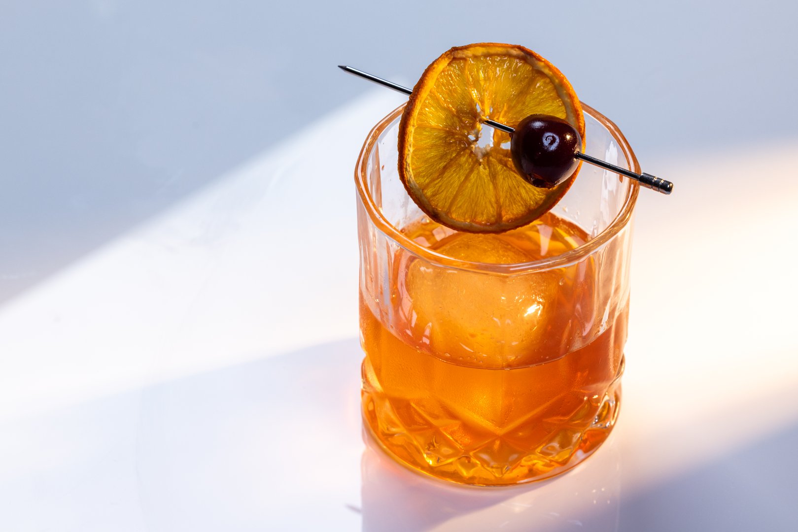 Close up of a manhattan cocktail served with one large ice cube on a reflective white surface with dual lighting tones and high contrast lights adding drama.