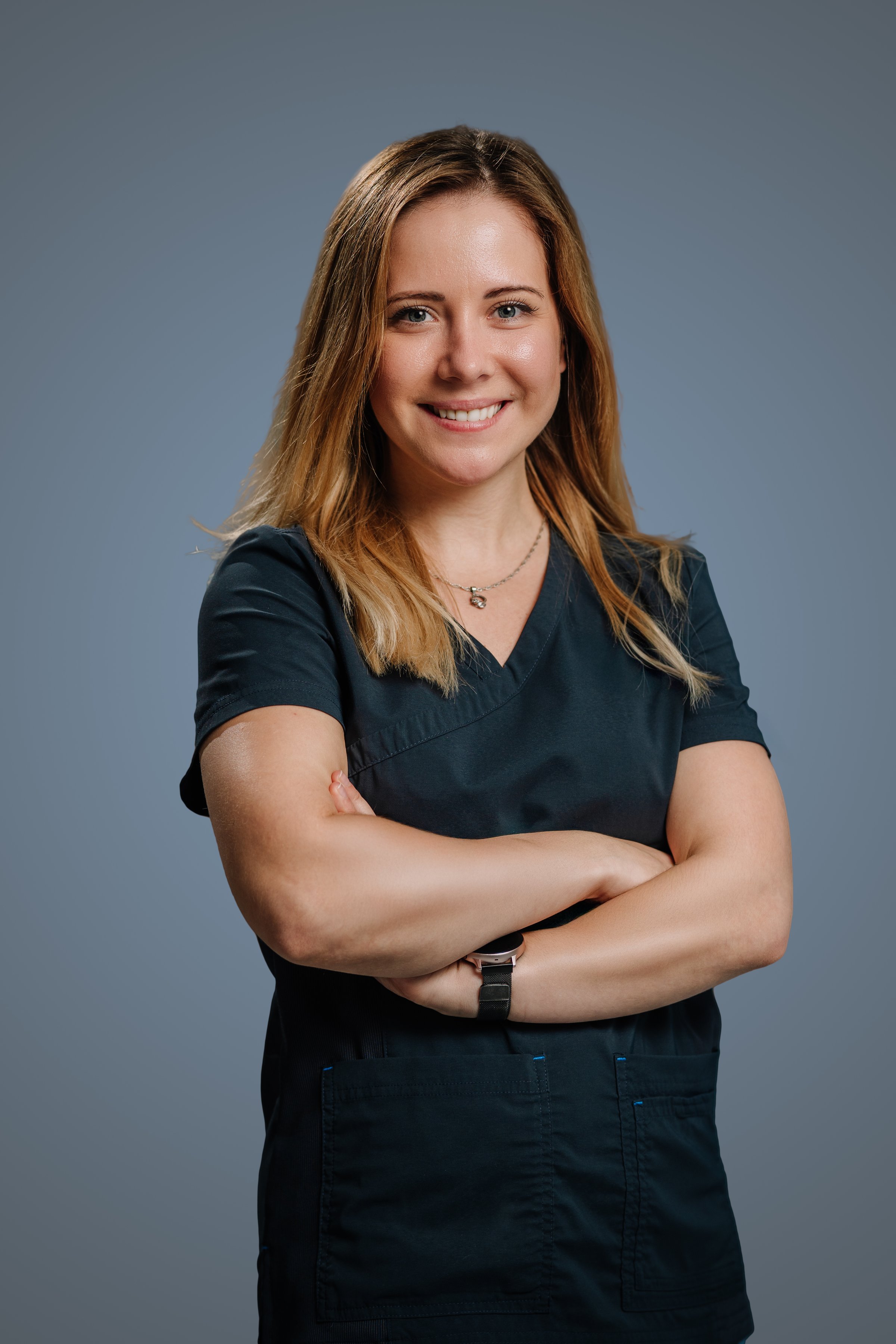 Confident woman in medical scrubs standing with arms crossed, smiling in a professional studio setting with a neutral background.