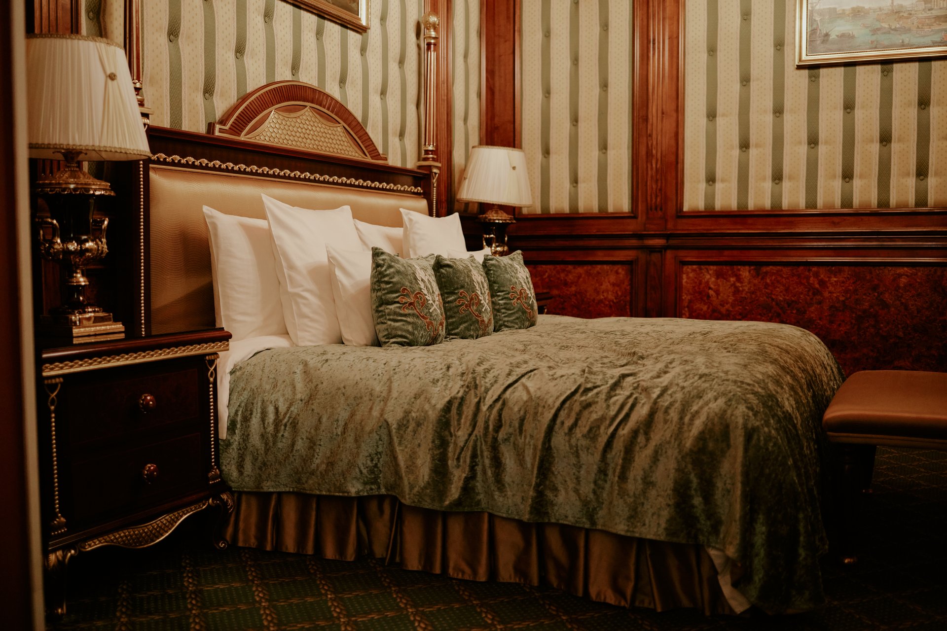 Hotel room featuring large bed with multiple pillows and decorative cushions, classic wooden headboard, two bedside tables with lamps, patterned wallpaper and carpet