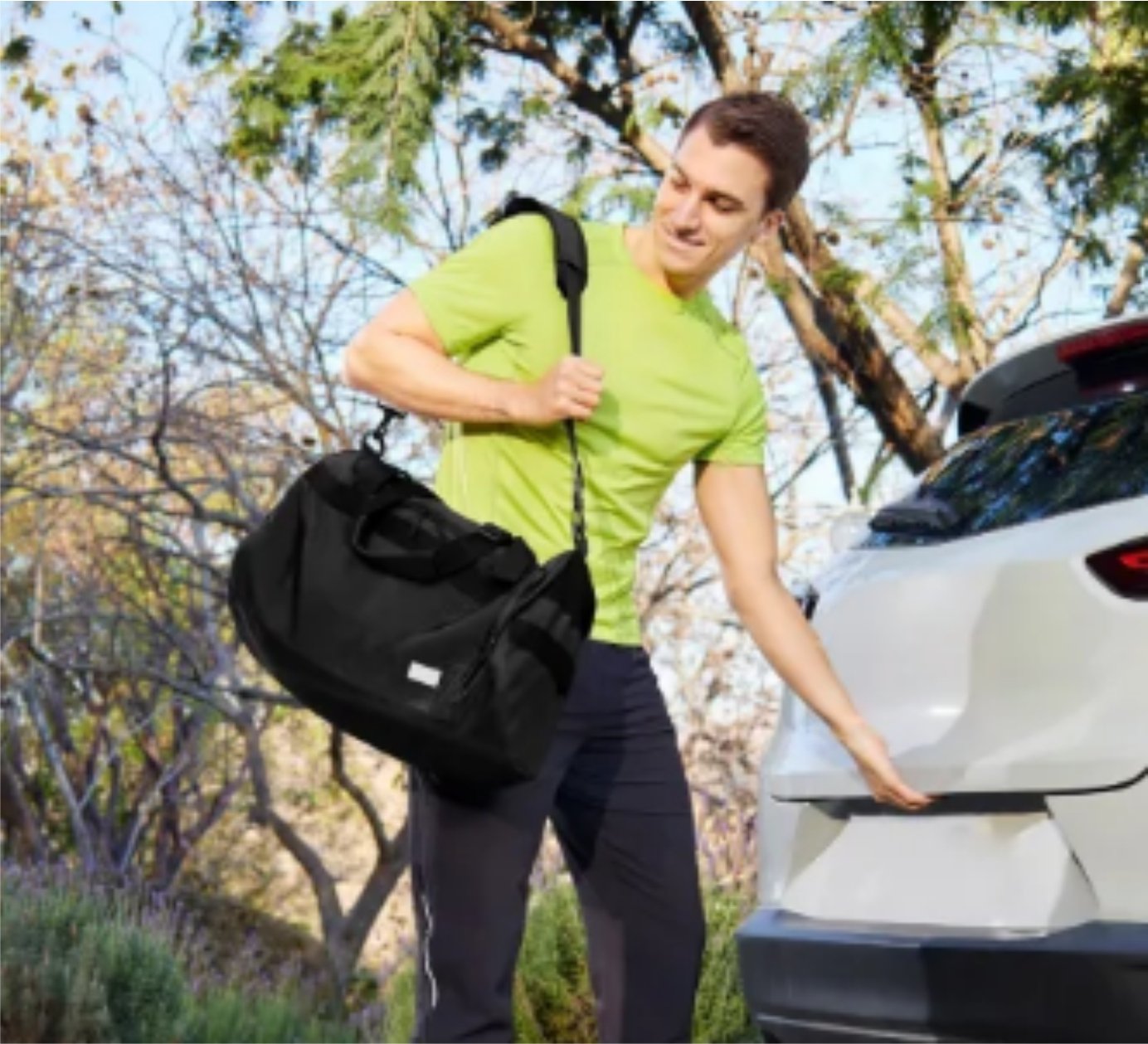 Man in a bright green shirt holding a black duffel bag and opening the trunk of a white car in a wooded area.