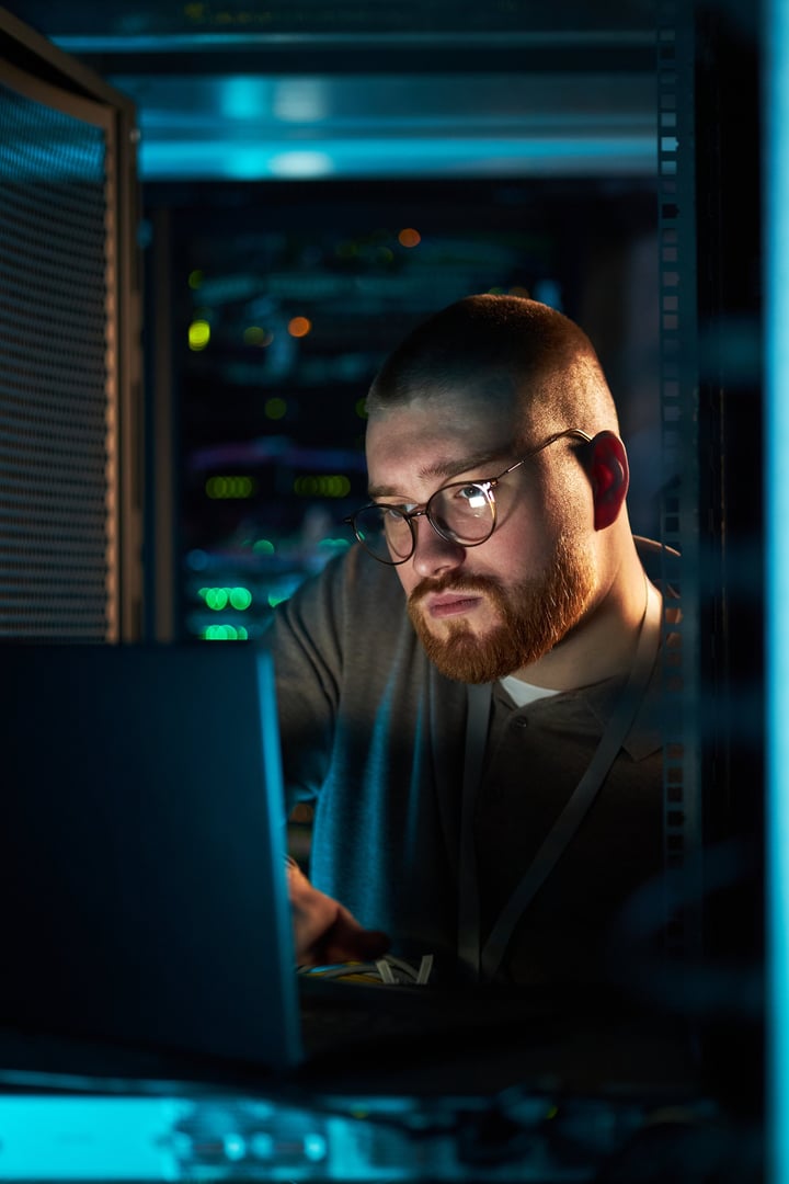 Person with beard wearing glasses working on laptop in data center surrounded by server racks creating sense of concentration and focus in technological environment