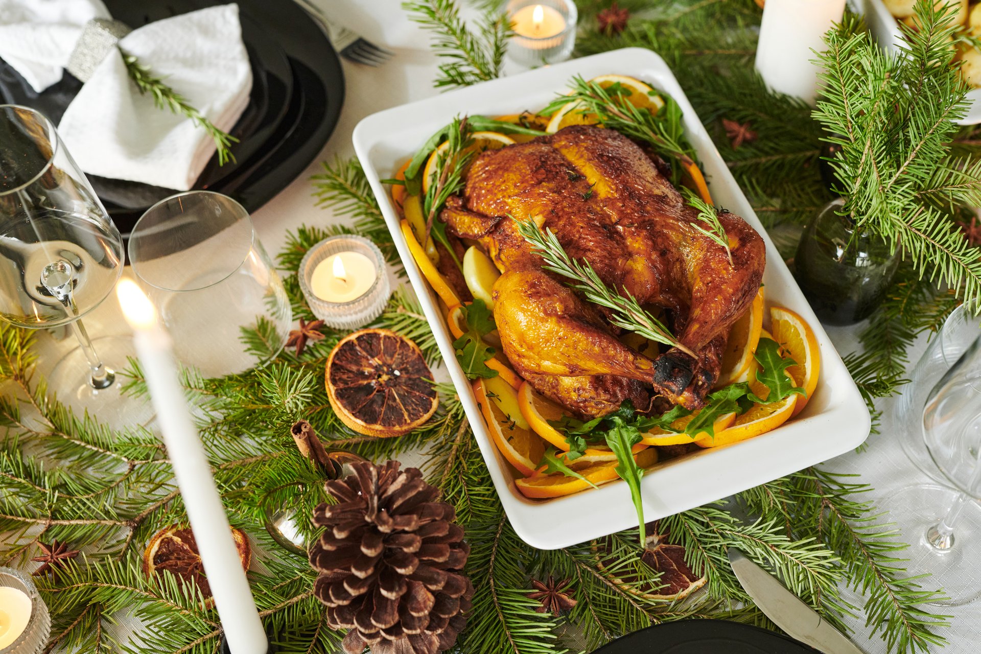 Whole roasted chicken garnished with rosemary and orange slices resting in baking dish surrounded by pine branches, candles, pine cone, wine glasses, Christmas holiday table setting