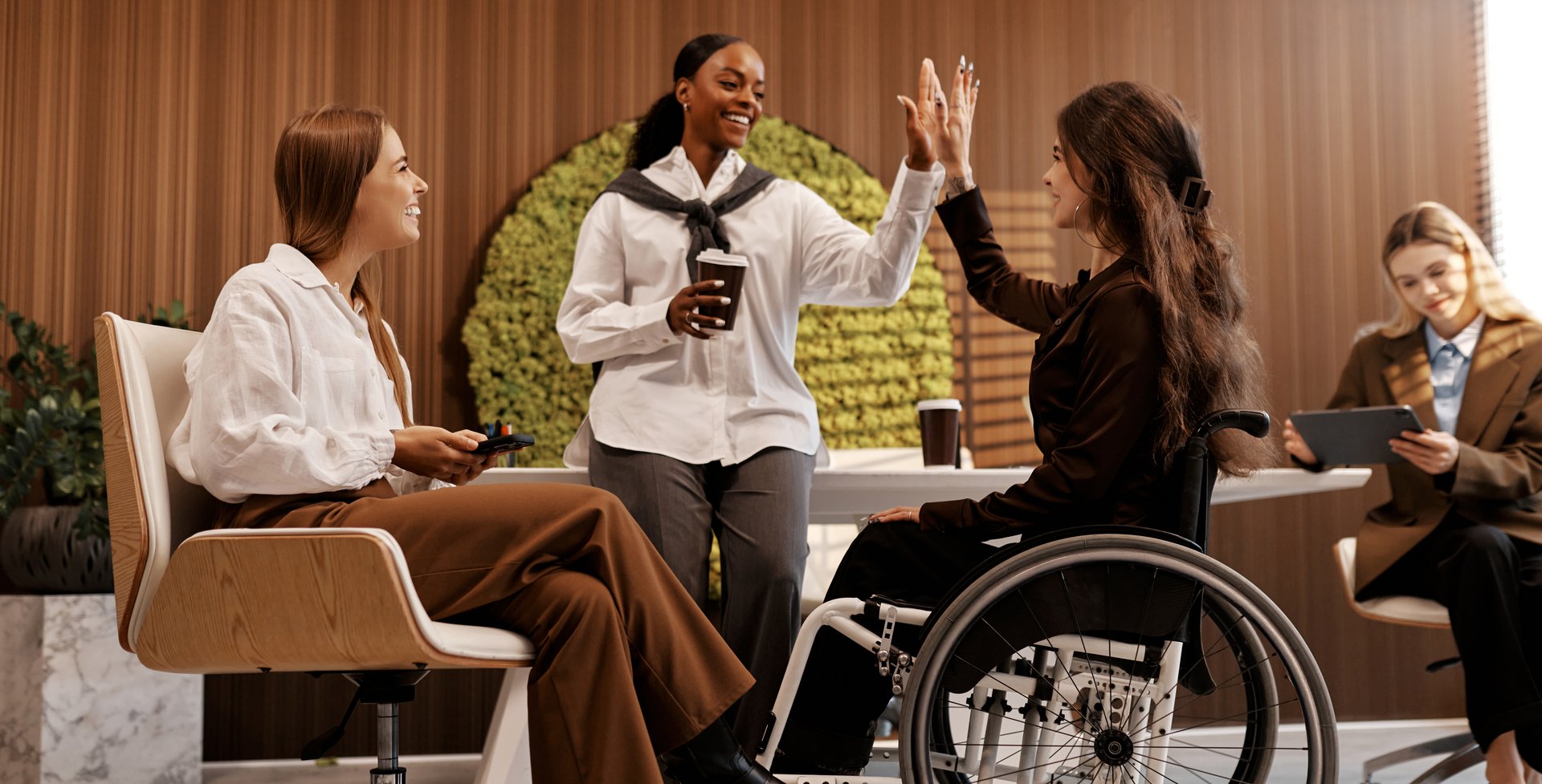 Diverse team of young women in office, including woman in wheelchair, giving high five and smiling. Workplace inclusivity, teamwork, support and celebration concept