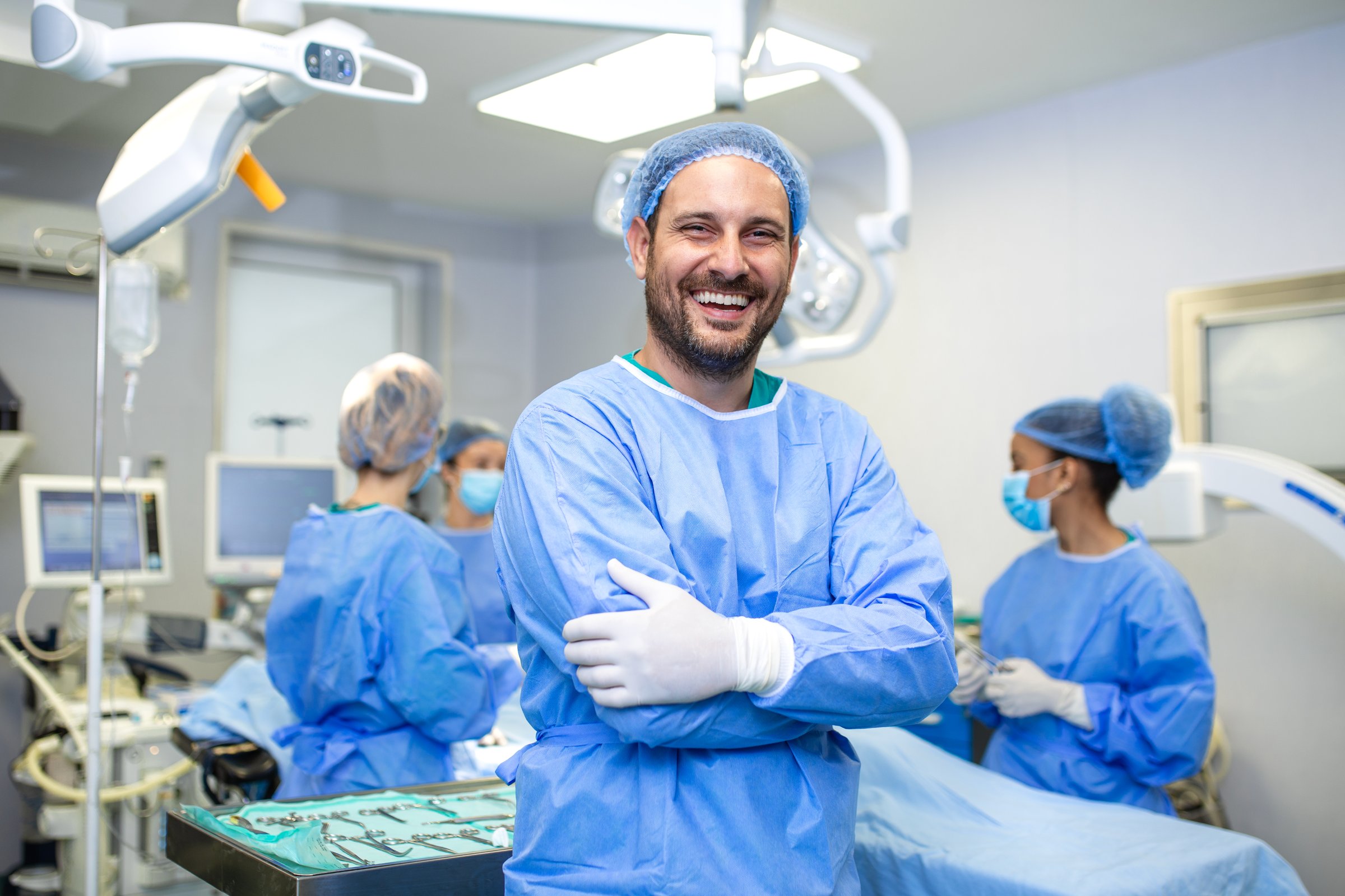 male surgeon stands with arms crossed and a big smile in an operating room while his team prepares in the background. Image conveys confidence, professionalism, and positivity in a medical environment