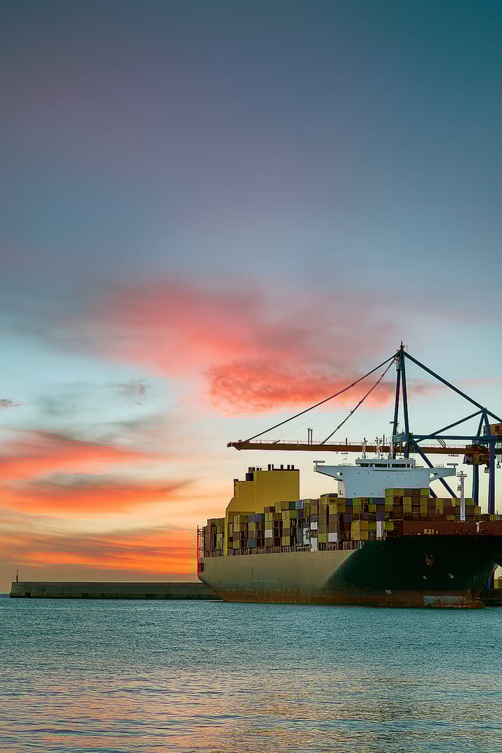 Minimalist view of a large cargo ship loaded with shipping containers docked at the harbor during sunset. The vibrant orange and pink clouds contrast with the calm sea, symbolizing maritime trade, global commerce, and the intersection of industry and natural beauty.