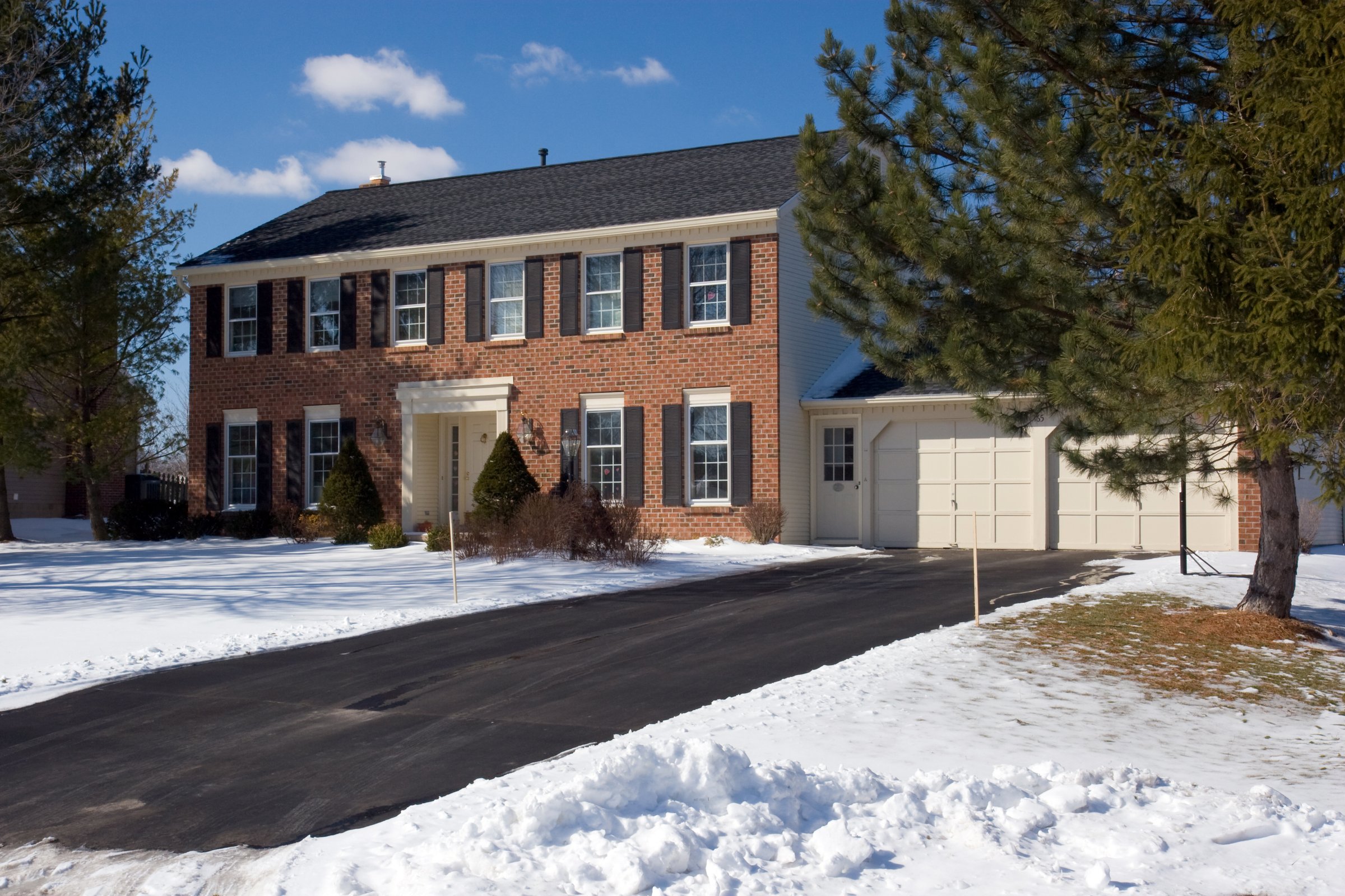 Classic red brick colonial house in the winter, with snow in the front yard, pine trees, a cleared black top driveway, and a bright, day time blue sky background.