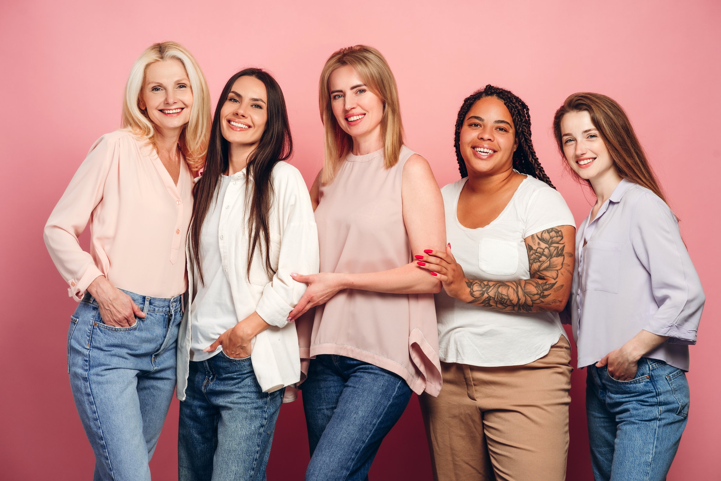 Five beautiful women of different ages and ethnicities are posing together on a pink background, showcasing diversity and female empowerment