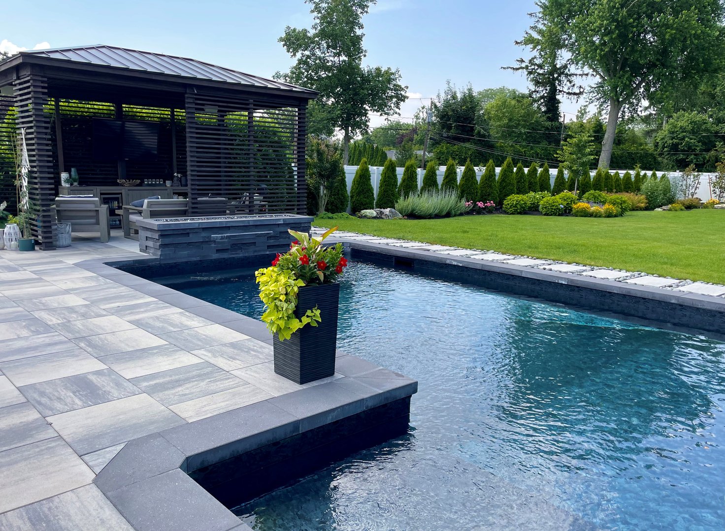 High-end, NY outdoor living space, a backyard patio with pergola, outdoor rug,  tequila bar, dining set, couch, television, pool with waterfall, umbrella, fire pit and stunning grey patio. Modern vibe. Surrounded by lush cottage style gardens, flowers, shrubs and trees.  Stunning entertainment space to be used all year round and to enjoy in the summer time.  The pool has a non traditional shape with a sundeck.