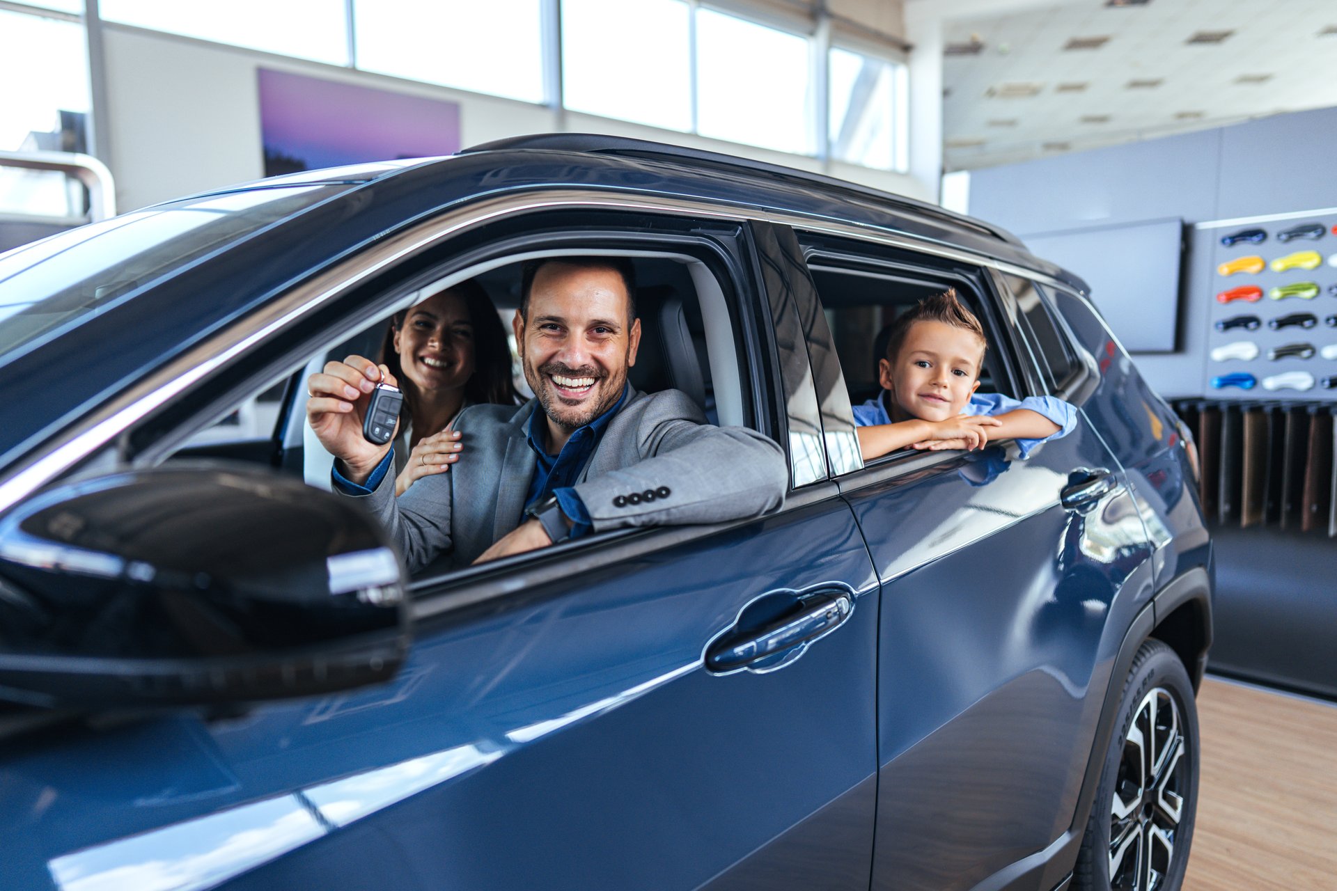 A joyful family sits in their new car at a dealership. The father holds the key, while the mother and child share the excitement of a new vehicle acquisition.