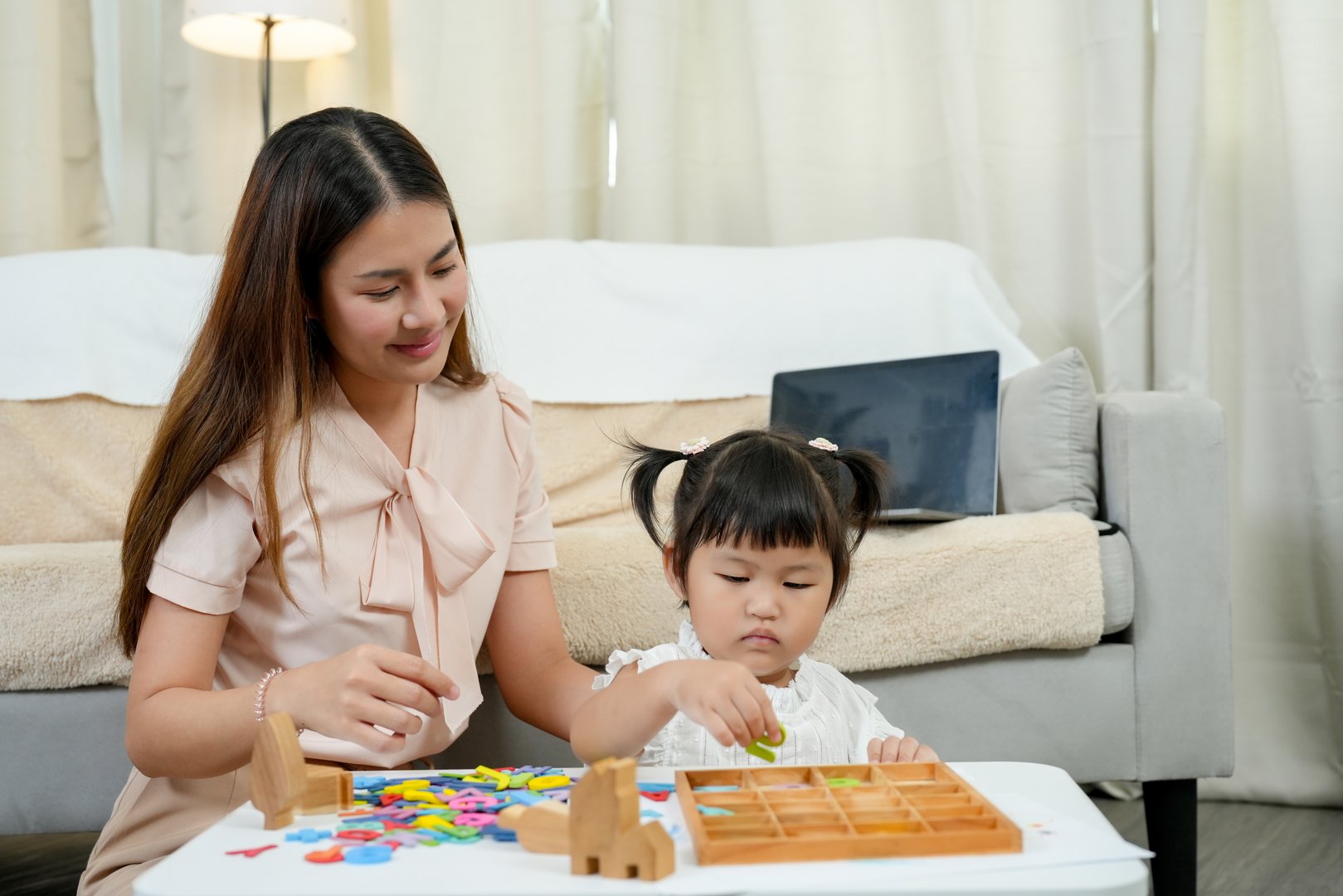 Asian preschool girl practicing problem-solving skills with wooden educational toys while female mentor encourages cognitive learning through structured play-based activities early childhood