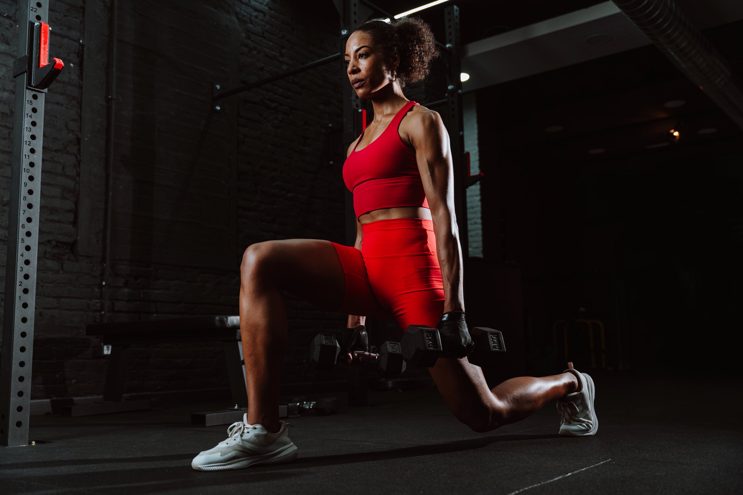 Cinematic image of a fit young woman in red sport outfit making workout in the gym. Woman doing lunges with dumbbells.