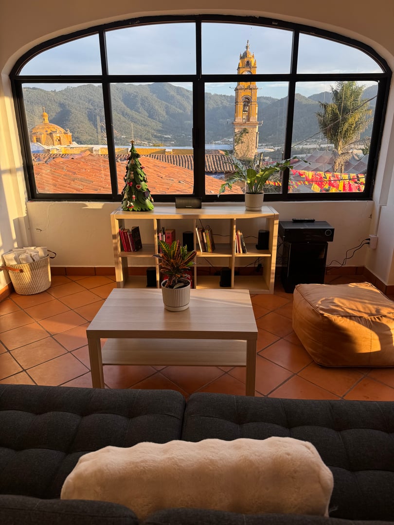 Cozy room with a large window view of a church tower and mountains, featuring a sofa, low table, and Christmas tree.
