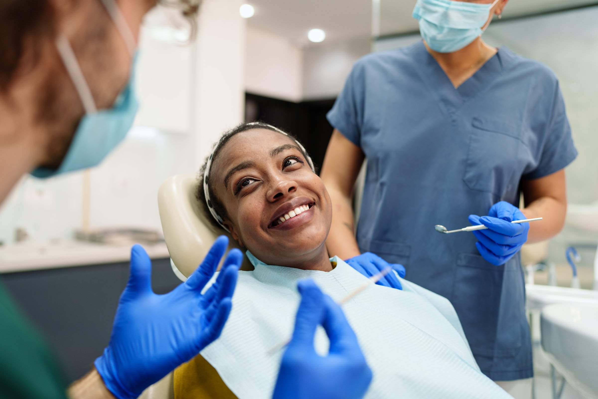 A Black woman smiles warmly while sitting in a dental chair. She is attended by a diverse dental team, including a male and a female, both wearing blue scrubs and gloves. The setting is bright and modern.