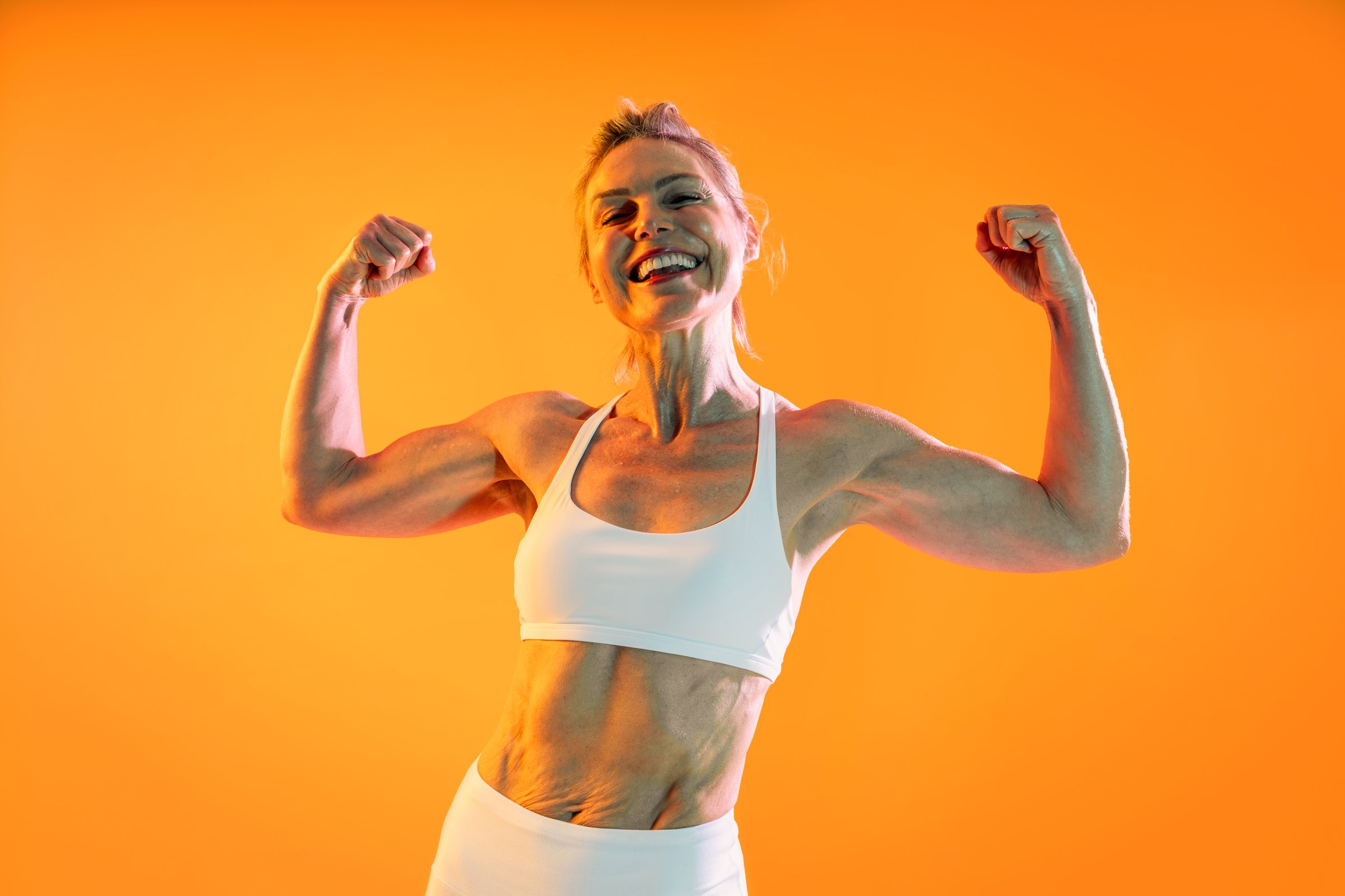 Beautiful mature sportive woman wearing sportswear doing workout - happy, mature woman in sportswear flexing biceps after a successful workout on an orange background, embodying strength and wellness