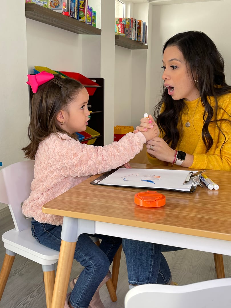 Child holds a marker, sitting at a table with a woman holding a clipboard; both are engaged in an activity.