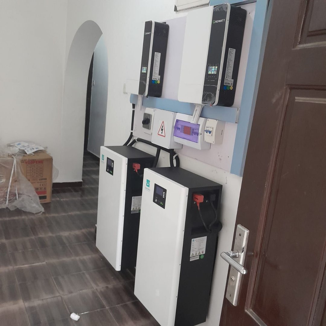 Wall-mounted inverters and batteries in a room with a tiled floor and a wooden door.