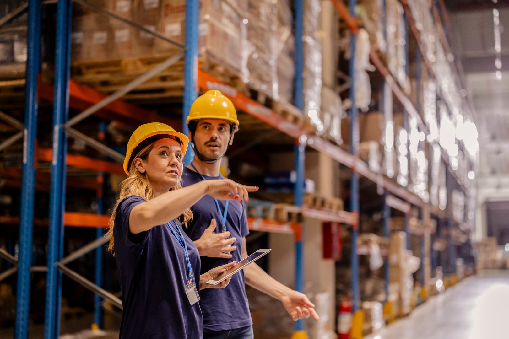 Two warehouse workers in safety gear discussing operations and reviewing inventory on a tablet in a warehouse aisle