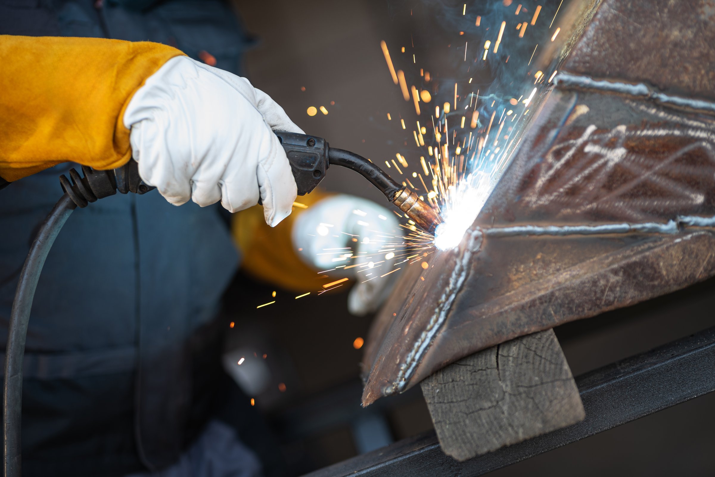 Welder wearing protective gloves is joining metal parts with a welding torch, creating a shower of sparks