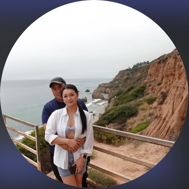 A couple poses in front of a scenic coastal cliff with ocean views, standing on a path with railings.