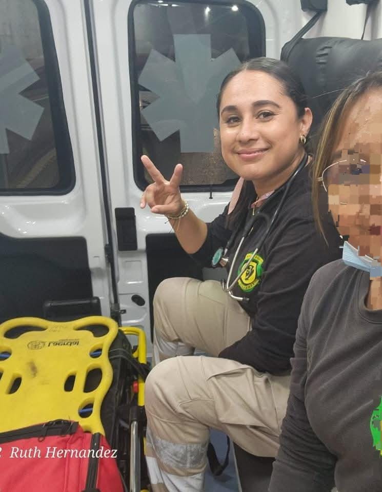 Two paramedics inside an ambulance, one giving a peace sign, with medical equipment visible in the background.