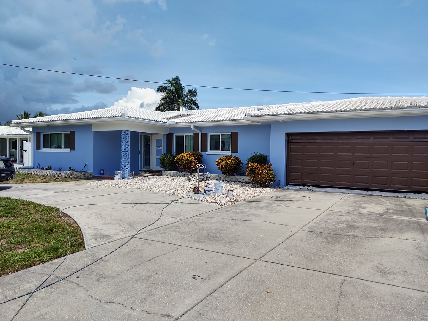 Single-story blue house with a white roof, brown garage door, and driveway. Palm trees and bushes are in the background.