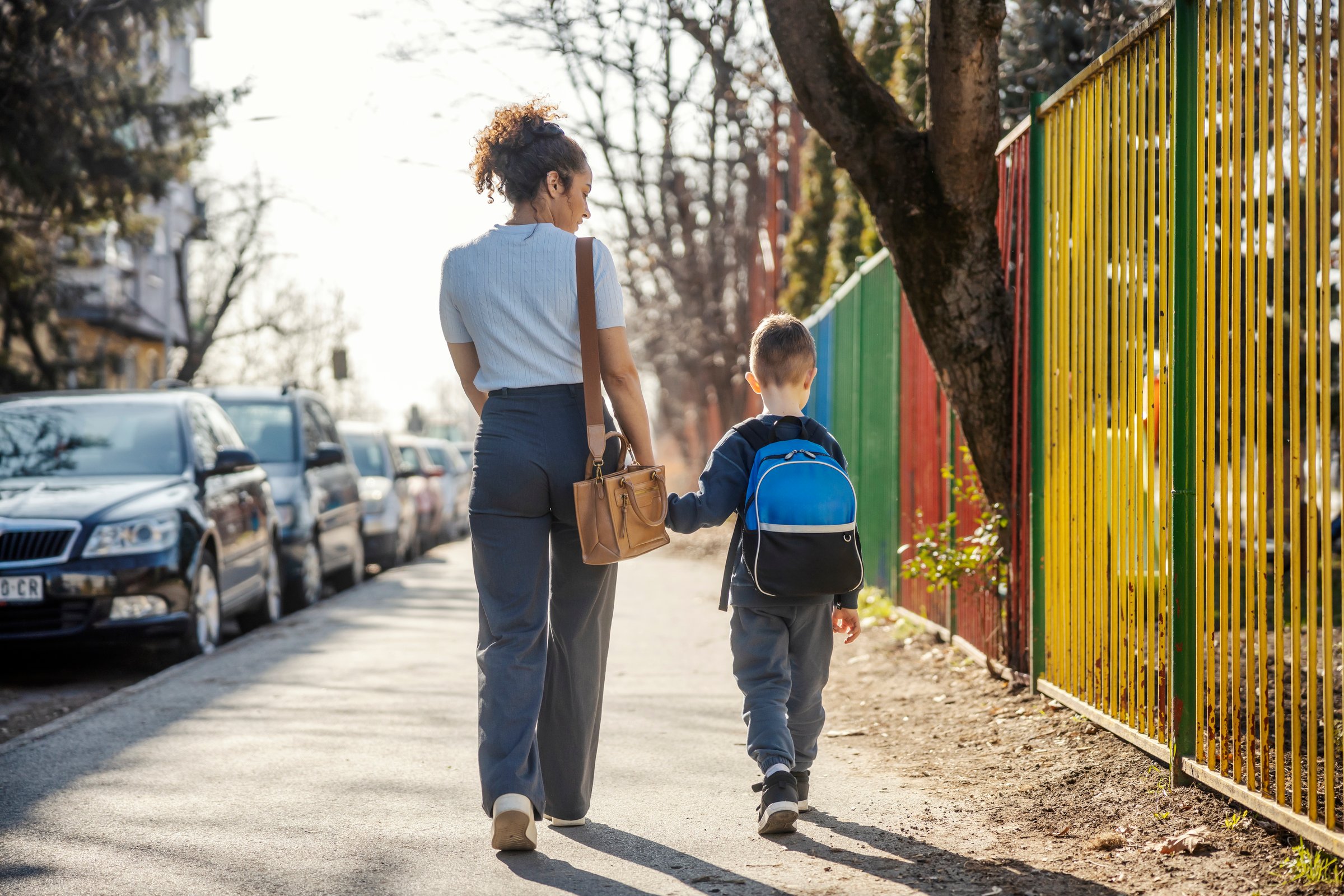 Back view of a young mother holding hands with her son and walking on a city street while going to the kindergarten.