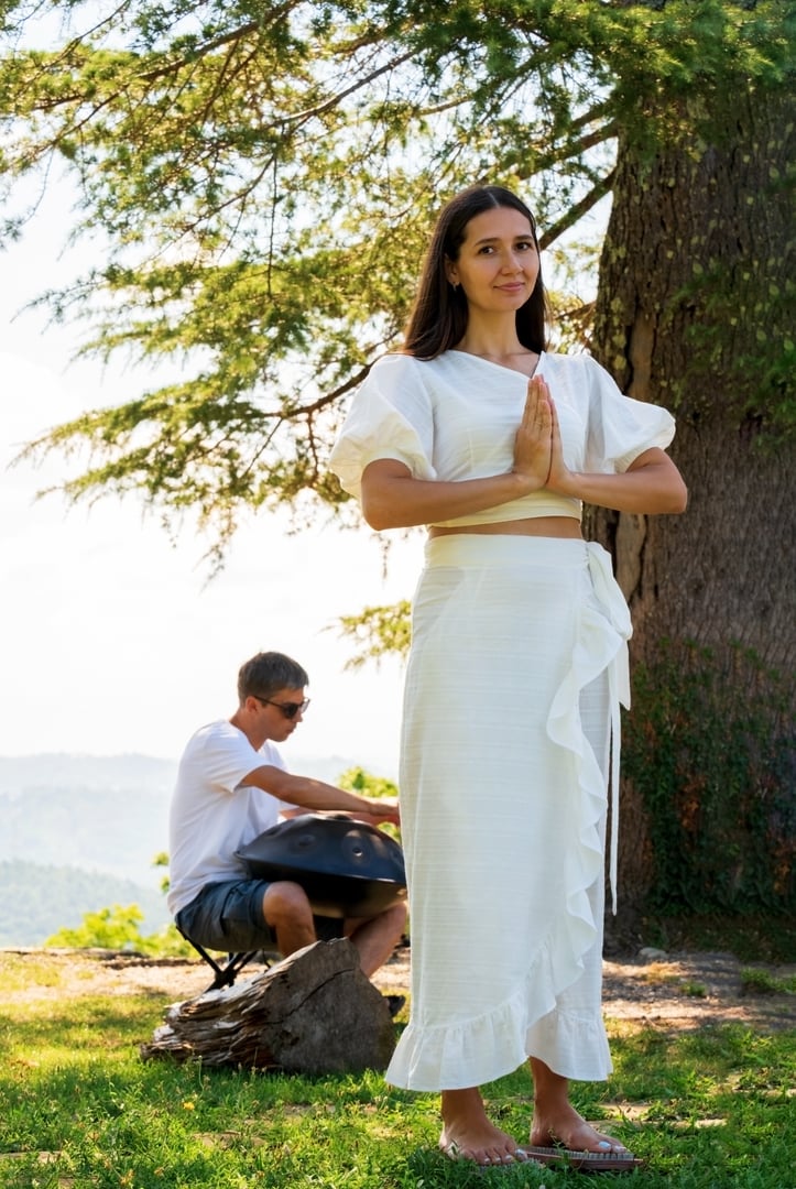 A young woman in a white dress is standing on nails, and a man is playing a handpan. Yoga retreat concept.