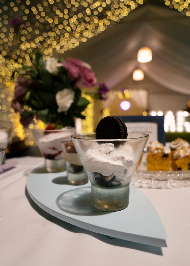 Catering service for a birthday. Closeup view of the sweet table with  a cookies and cream dessert in a cup of glass. The lights, floral arrangement and tent in the background