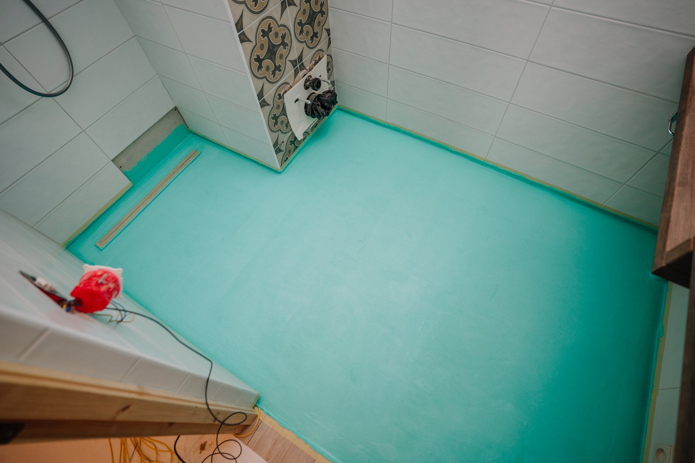 Freshly waterproofed bathroom floor with a blue membrane layer, tile walls, linear drain, and visible electrical wiring during renovation.