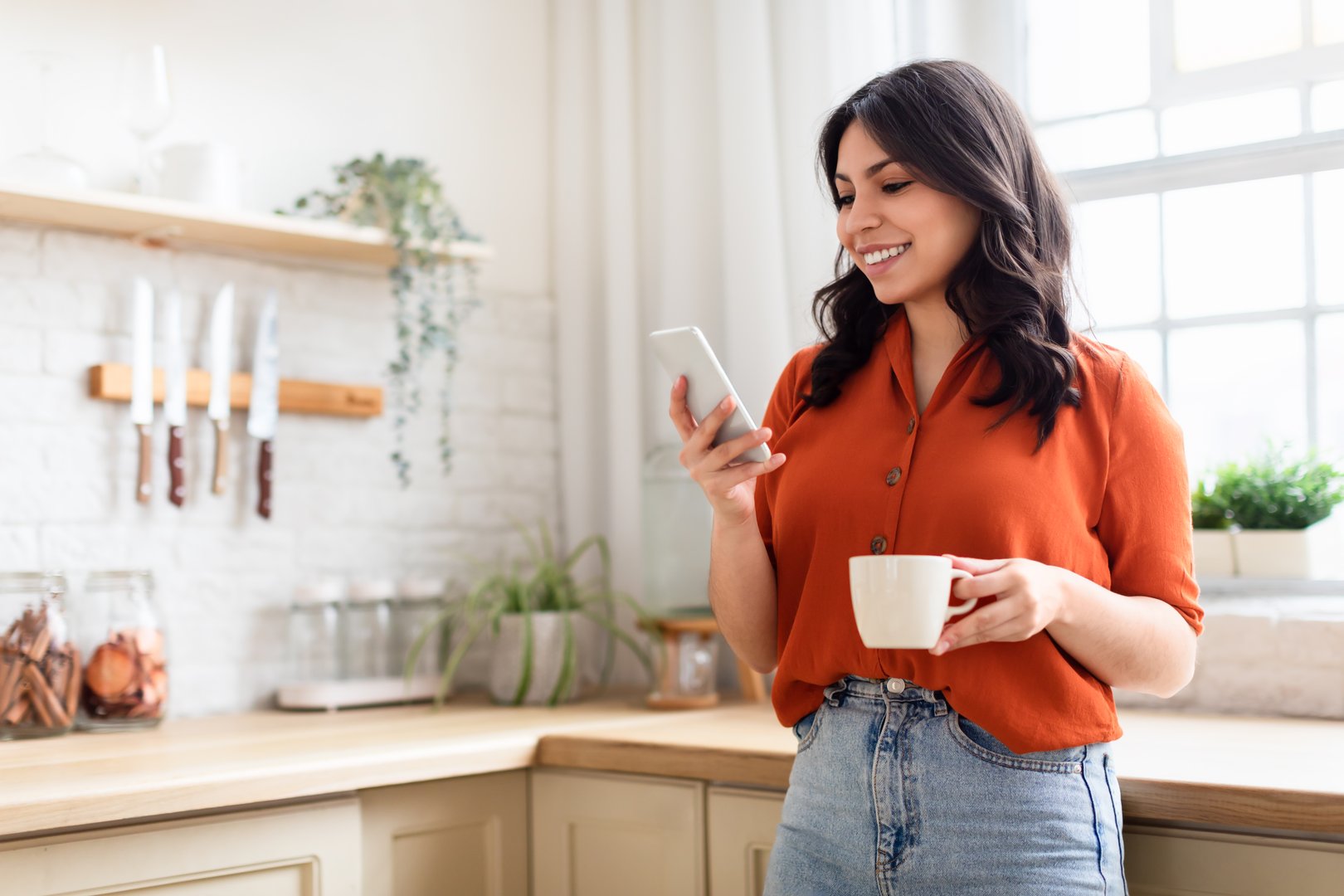 Smiling young woman enjoying her coffee while using her smartphone in a homey kitchen setting, copy space