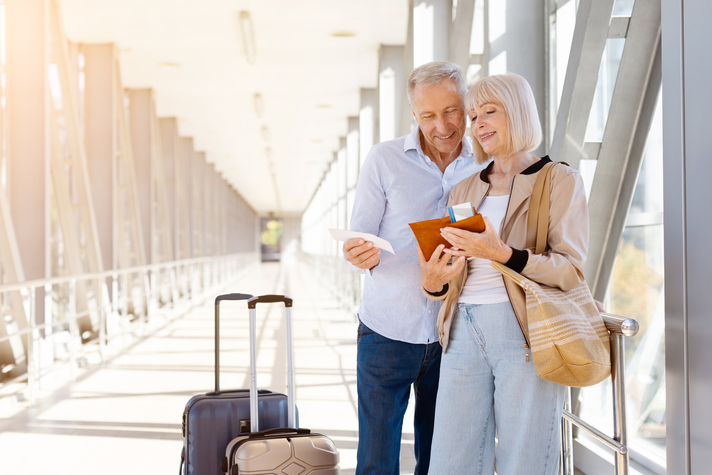 Smiling mature couple standing at airport corridor with luggage, reading passports and tickets together. Family lifestyle, travel preparation, and retirement joy.