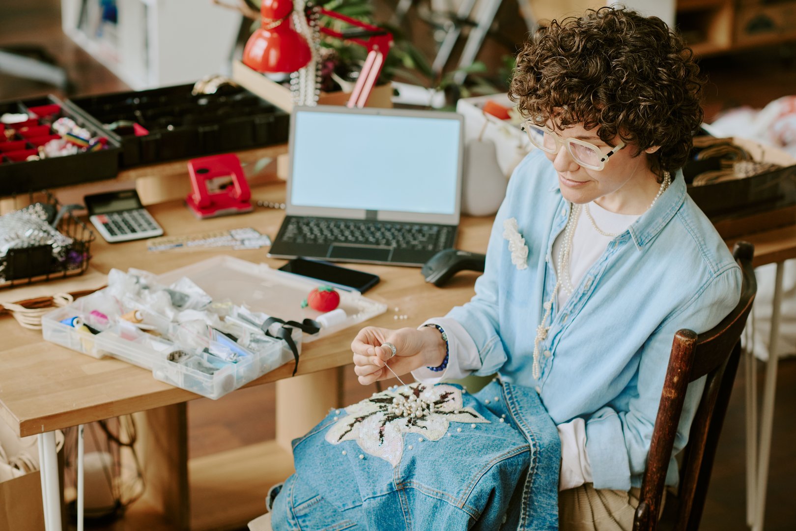 Person involved in detailed embroidery work on jacket at home office desk, surrounded by accessories and materials, focusing on intricate patterns without looking straight at camera