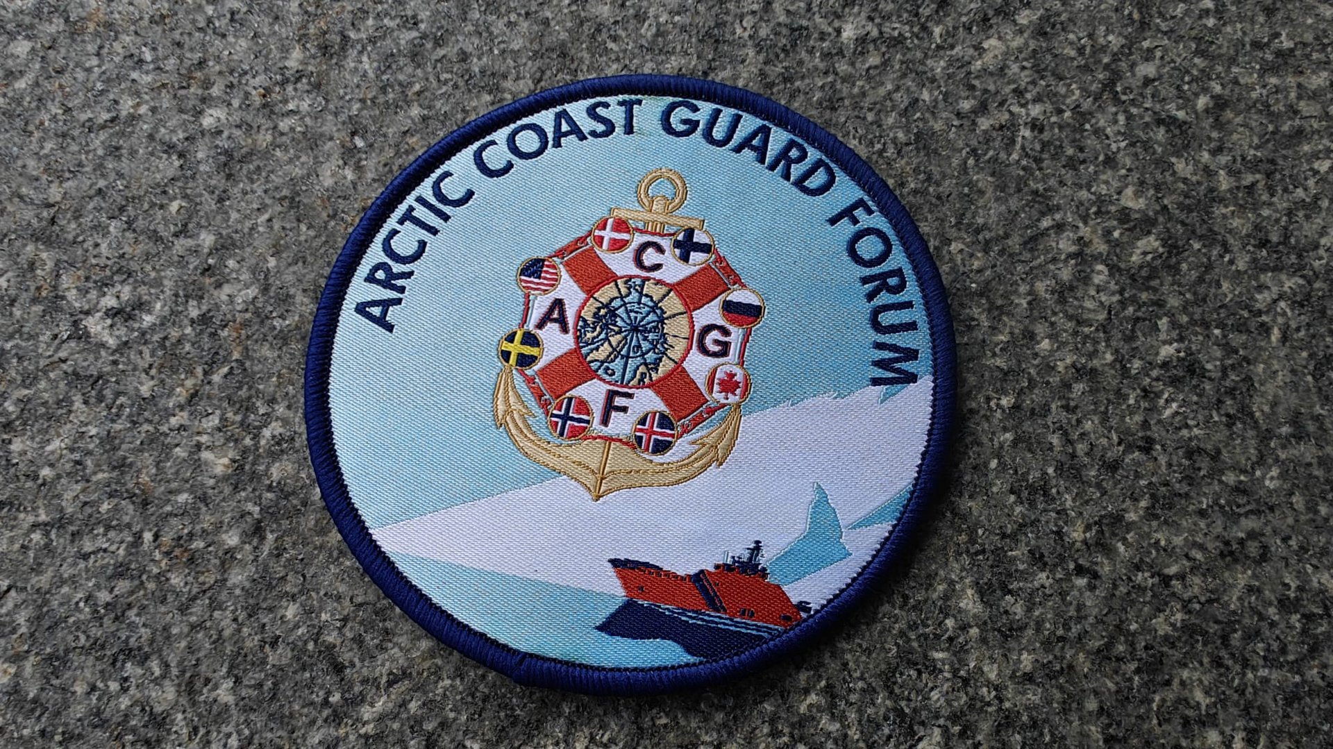Arctic Coast Guard Forum patch with a ship, flags, and compass design on a blue and white background, placed on gray stone.