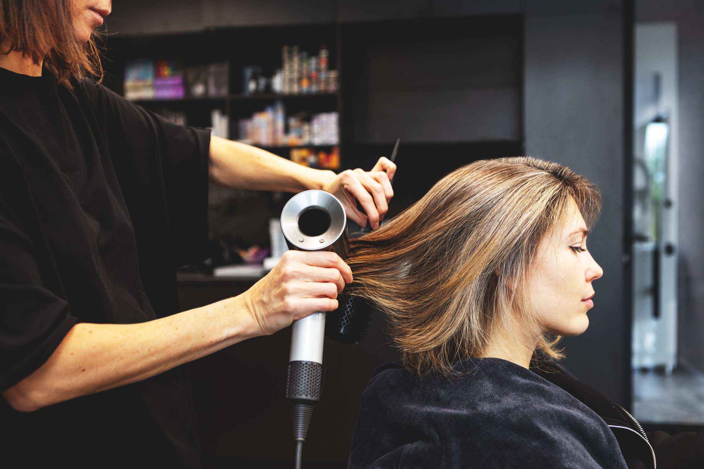 Hairstylist blow drying womans hair with brush and blow dryer, Side view hairstyling process in modern beauty studio with dark interior, professional styling