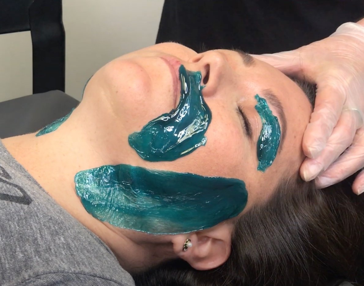 Person receiving a facial treatment with teal gel applied to the chin, cheeks, and forehead, gloved hands touching the face.